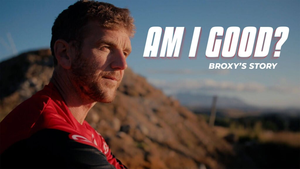 Am I Good? Broxy's story. A man looks passed the camera with the setting sun lighting his face. He sits on a motocross jump.